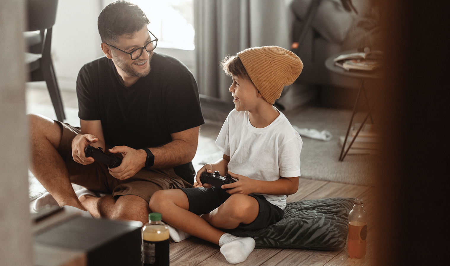 a man with a young boy, sitting on a floor in a living room, holding Play Station controllers, smiling to each other 